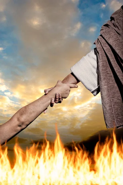 Hand of Jesus Christ giving helping someone to get out of the flames of hell. Background of fire and cloudy sky.