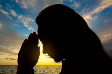 Woman's face backlit with hands in prayer on a sunset background. clipart