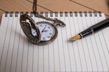 elegant black fountain pen with gold details and patinated bronze pocket watch with decorative engraving of antique locomotive on lined spiral-bound notebook with thick covers on wooden background clipart