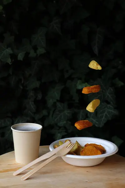 Takeaway - concept - with falling food - disposable wooden cutlery and chinet disposable bowl with fried food and disposable cup on wooden table, dark background, space for text, eco friendly