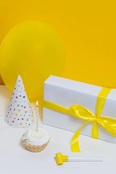 Serving size dessert topped with white frosting with one burning candle, gift decorated with yellow ribbon and cone cap on the table. Happy Birthday card design concept.