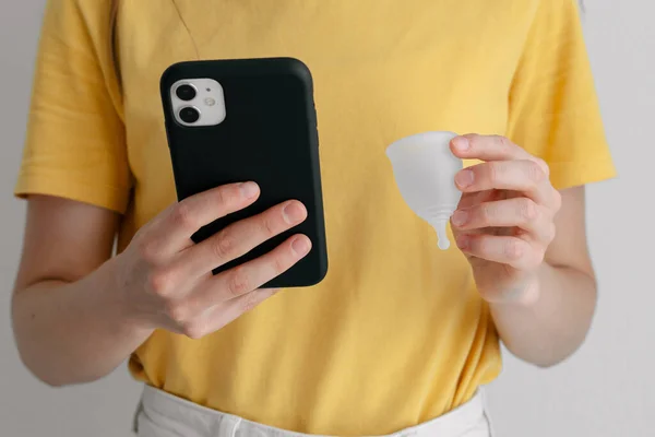 young woman with menstrual cup and phone reads instructions close-up. hypoallergenic silicone cup for menstrual blood