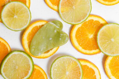 Yeşil guaj masaj aleti, su damlacıklarıyla kaplı beyaz bir zemin üzerinde turuncu ve limon dilimlerinin üzerinde bulunur. Tasarımınız için arkaplan