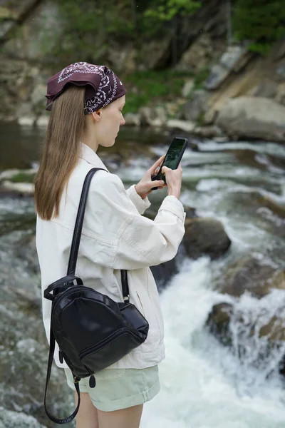 A young wearing a white denim jacket and pants is filming a rapid river and a cliff on her phone. The concept of oneness with nature, solo travel