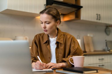 Genç bir kadın masada oturuyor ve mutfakta dizüstü bilgisayar kullanarak ders çalışıyor. Uzaktan öğrenme ve çalışma kavramı