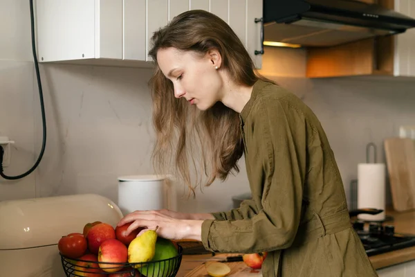 stock image A young pretty woman in a khaki dress is choosing fruits in a basket in a modern kitchen. Grocery shopping and healthy eating concept