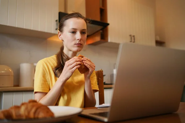 A young woman is eating a croissant and looking at a laptop while sitting at the table in her kitchen. The concept of remote work, entertaining videos for distance learning