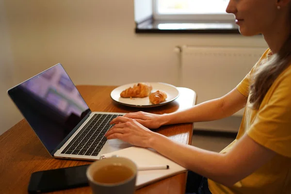 Young woman typing text on laptop with black screen while sitting at table with food and notebook at home in kitchen. The concept of remote work