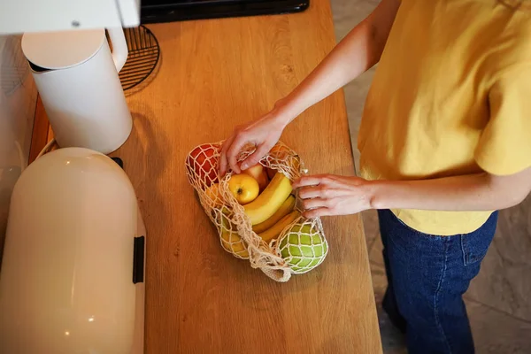 A woman in a yellow T-shirt unpacks an ecological mesh bag with organic products in her kitchen. The concept of reusable packaging