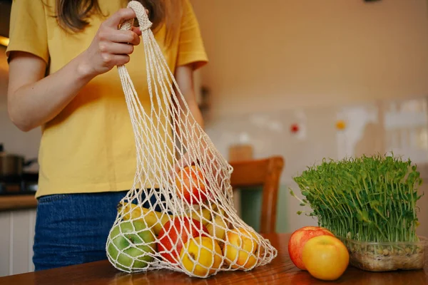 A woman in a yellow T-shirt takes products from an ecological bag to the table. Concept of organic products, healthy habits and reusable packaging