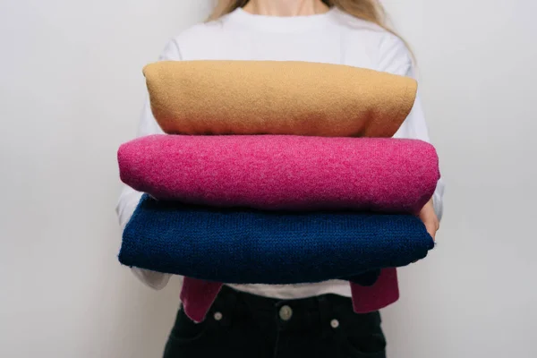 A young woman holds three warm woolen sweaters of different colors in front of her on a white isolated background. The concept of changing winter clothes