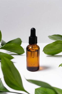 Glass brown reusable bottle for natural serum on white isolated background with green plant leaves. Cosmetic product presentation or image for your design
