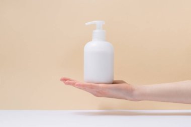 A female hand holds a blank white cream bottle on a beige background with a shadow. Image for your design or mockup