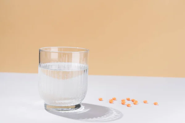 Horizontal image of a transparent glass with pills on a beige background. Vitamins, bad and medical concept.