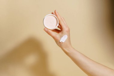 Bej izole arka planda gölgesi olan nemlendirici leke dolu bir kavanoz tutan kadın eli. Yüz bakımı kavramı, kırışıklıkları gidermek.