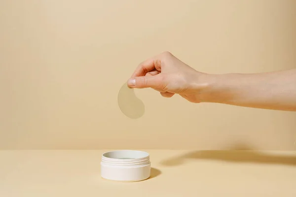 stock image Female hand holding sample of green algae extract eye patch over white jar of product on beige isolated background. The concept of natural means for moisturizing, from dark circles under the eyes, from wrinkles