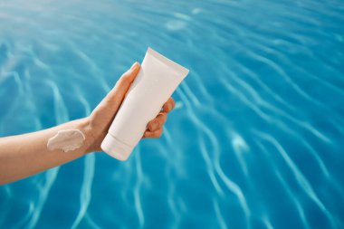 Mavi havuz suyunun arka planında beyaz bir güneş kremi tüpü tutan lekeli bir kadın. Yüz için nemlendirici kozmetik kavramı, yaz tatili, güzellik, kaplıca ve estetik