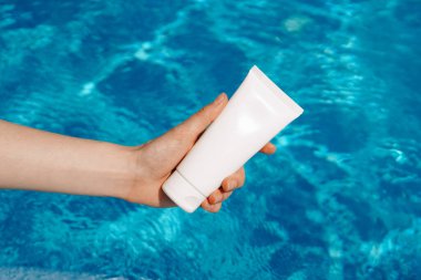 Kadınların narin elleri havuzun arka planında beyaz bir nemlendirici ya da güneş kremi tutuyor. Koruyucu cilt, güzellik ve kendine bakım kavramı