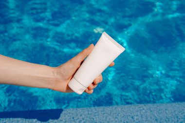 Genç bir kadının eli, mavi suyun arka planında gündüz güneşinin aydınlattığı beyaz bir krem tüpünü tutuyor. Güneş koruması ve nemlendirici cilt kozmetiği, güzellik, estetik