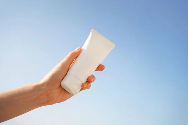 Female hand holding a white tube of sunscreen mockup on a blue sky background. Spa concept, moisturizing and skin regeneration after long exposure to the sun and exposure to uva and uvb rays