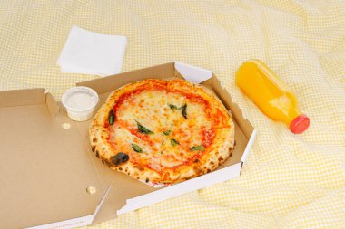 İçinde sos ve portakal suyu olan bir kutuda pizza ve sarı bir piknik paspasında cam bir maket şişesi. Fast food, dağıtım ve açık hava eğlencesi konsepti.