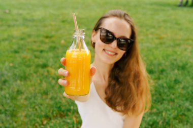 Park arka planında gözlük takan genç bir kadının tuttuğu portakal suyu ya da multifruit suyu olan cam bir mockap şişesine odaklan. Müşteri tarafından kullanılan meyve suyu ve nektarin paketleme ve satma kavramı