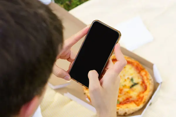 A young man sit in a black mock-up phone on the background of a pizza in a box. Fast food delivery concept. Image for your design