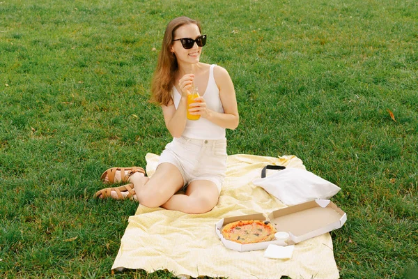 A young stylishly dressed woman drinks orange or peach juice from a glass bottle and eats pizza while sitting on a picnic. Concept of outdoor recreation, use and packaging of fruit juices or drinks