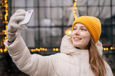 Genç ve çekici bir kadın telefonda, parlak çelenklerin arkasında selfie çekiyor.