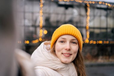 Sarı şapkalı çekici bir kadın kışın sokakta bir çelengin arka planında selfie çekiyor. Tasarımın için resim