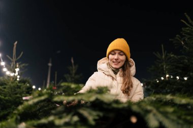 Sarı şapkalı ve kışlık giysili gülümseyen genç bir kadın çelenklerin arasından bir Noel ağacının dallarını seçiyor. Yeni yıl tatili ve mucizeleri konsepti