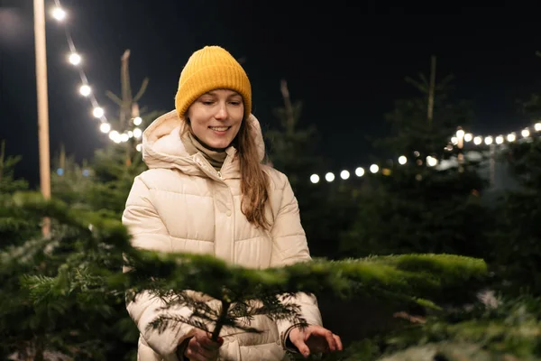 A young smiling woman chooses Christmas tree branches to decorate the house for the New Year holidays against the background of bright lights.