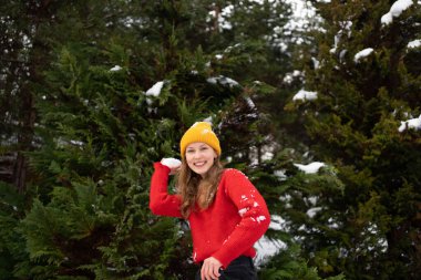 Genç, gülümseyen kadın. Kırmızı örülmüş bir kazak, yeşil Noel ağaçlarının arka planına bir kartopu fırlatacak. Kış eğlencesi, eğlence ve tatil aktiviteleri kavramı