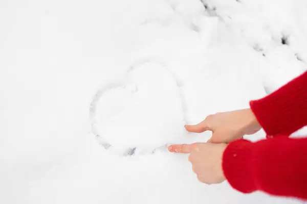 The hands of a young woman in a red sweater draw a heart on white snow. Concept of winter activities and fun