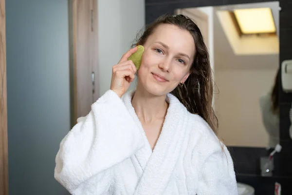 Portrait of a young woman in a bathrobe with a green gouache massager on a mirror background. The concept of face care, lifting and beauty procedures