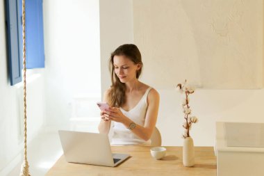 Oturma odasında dizüstü bilgisayarı olan genç bir kadın ahşap bir masada otururken telefonla konuşuyor. Uzaktan çalışma, çevrimiçi kurslar ve eğitim kavramı