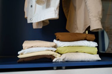 Mavi bir dolabın rafındaki üst üste yığılmış giysiler ve askılarda asılı gömlekler. Küçük bir gardırop deposu, oda konseptini düzenliyor.