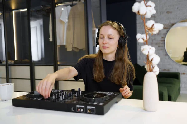A young woman mixes tracks on a DJ controller for a set while sitting in a room. The concept of hobby and remote work, preparation for the performance and online broadcasting