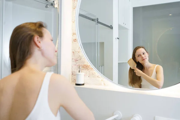 A young woman combs her shiny hair with a wooden comb while looking in the mirror in the bathroom. Self care concept, daily routine and hair cosmetics