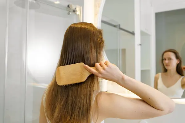 Young woman combs her shiny hair with a wooden comb, rear view. Concept of daily body care routine, hair cosmetics