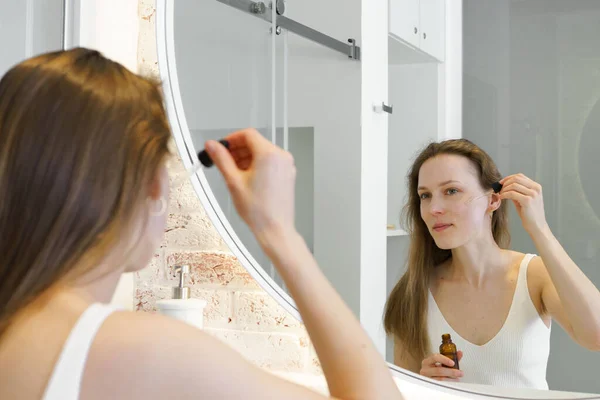 A young woman looks in the mirror and applies serum with a pipette to her face in the bathroom. Concept of skin care, cosmetic beauty products