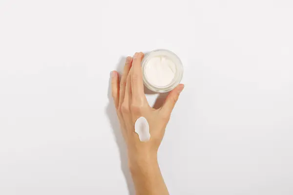female fragile hand with cream smear holding glass jar of face cream on white isolated background. concept of beauty products, skin care, cosmetology