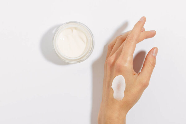 Fragile female hand with smear of cream and glass jar of facial moisturizer on white isolated background. Concept of skin care cosmetics, beauty products and cream sample
