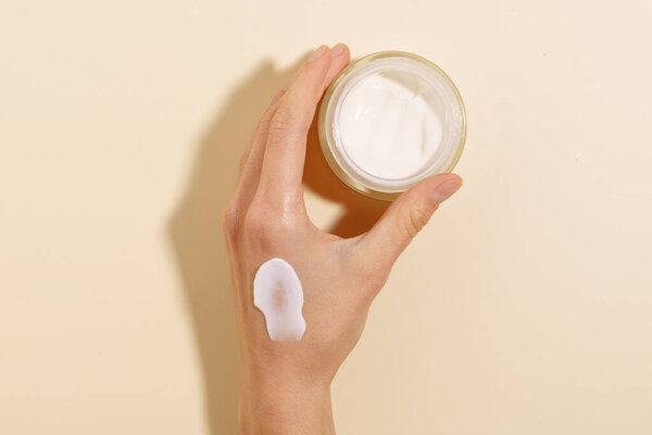 close-up of a female hand with a smear of cream holding a glass jar of moisturizer on a beige isolated background. concept of skin care and nutrition, beauty products