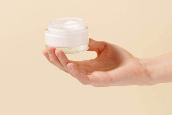 Close-up of fragile female hand holding glass jar of nourishing face cream on beige isolated background. Concept of beauty products, skin care, cosmetics and cosmetology