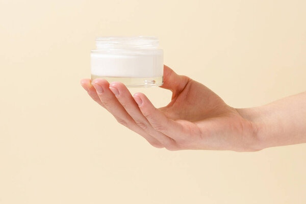 Female hand holding glass jar of facial moisturizer on beige isolated background. Concept of beauty products, skin care, cosmetics and cosmetology