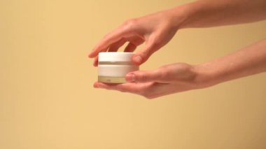 Kadın elleri cam maket kavanozunu açar ve bej kaplama arka planda cilde krem sürer. Güzellik, cilt bakımı, nemlendirici ve beslenme kavramı