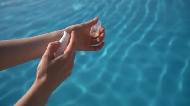 女性の手は白い瓶を保持し 青いプール水の背景に血清や油を塗ります 太陽からの夏の保護の概念 保湿および皮の滑らか — ストック動画