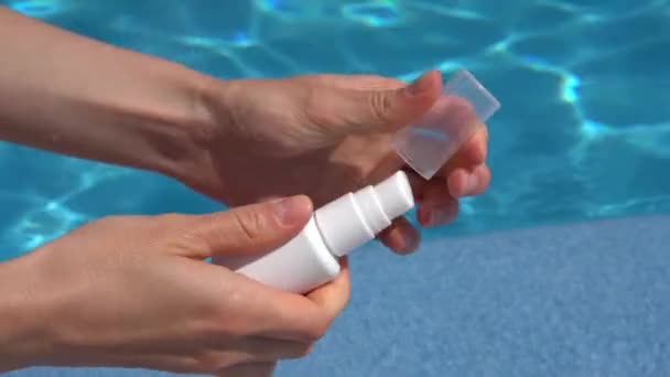 女性の手はディスペンサーが付いている皮の血清の白い瓶を開き 昼間の太陽によって照らされるプールの青い水の背景の手に適用します — ストック動画