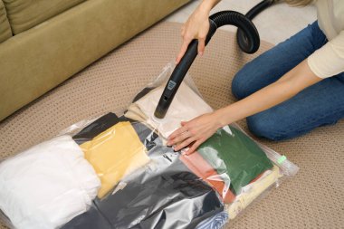 Genç bir kadın vakum torbasından havayı çekmek için elektrik süpürgesi kullanıyor ve yerde otururken dolapta giysi ve kompakt depolama için giysiler var. Yer tutma kavramı, sıkıştırılmış paketleme ve mühürleme